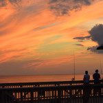 Two people fishing on a pier at sunset.