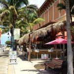 A sidewalk with palm trees and people sitting at tables.